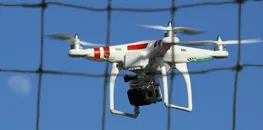 White drone with red stripes and an attached camera flying close to a net barrier against a clear blue sky