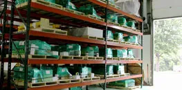 Industrial warehouse shelving filled with organized boxes, containers, and supplies, with an open entrance showing a glimpse of the outdoors.