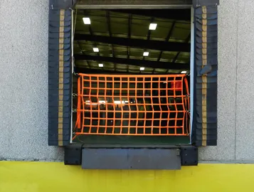 Orange safety net installed at a warehouse loading dock with black protective buffers, set against a bright yellow floor and dark interior.