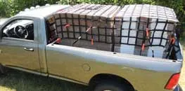 Pickup truck with its bed secured by a large orange and black cargo net, parked outdoors with greenery in the background.
